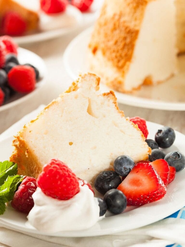A slice of fluffy angel food cake topped with fresh raspberries, blueberries, and sliced strawberries, accompanied by a dollop of whipped cream and a sprig of mint, served on a white plate. Other plates with cake and berries are in the background.