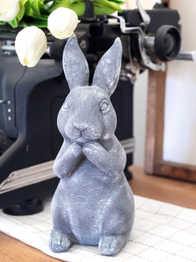 A gray ceramic rabbit figurine sits with its paws up to its mouth on a checkered cloth—an adorable touch for Easter decoration ideas to make. White tulips and a vintage typewriter peek out in the background.
