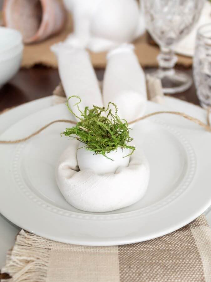 A white plate holds a rolled white napkin shaped like bunny ears, with an egg in the center topped with green moss—an easy Easter decoration idea to make for a rustic, spring-themed table setting on a beige placemat.