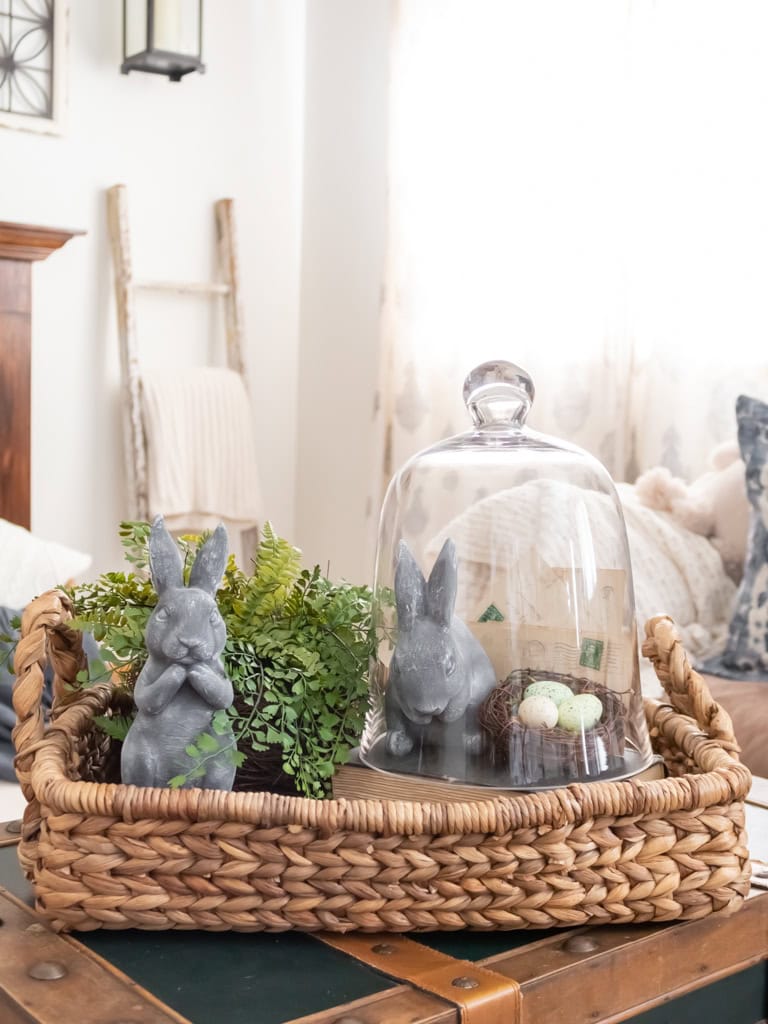 A woven tray holds two gray bunny figurines, a small green plant, and a glass cloche with one bunny and decorative eggs—perfect Easter decoration ideas to make for a cozy, light-filled living room.