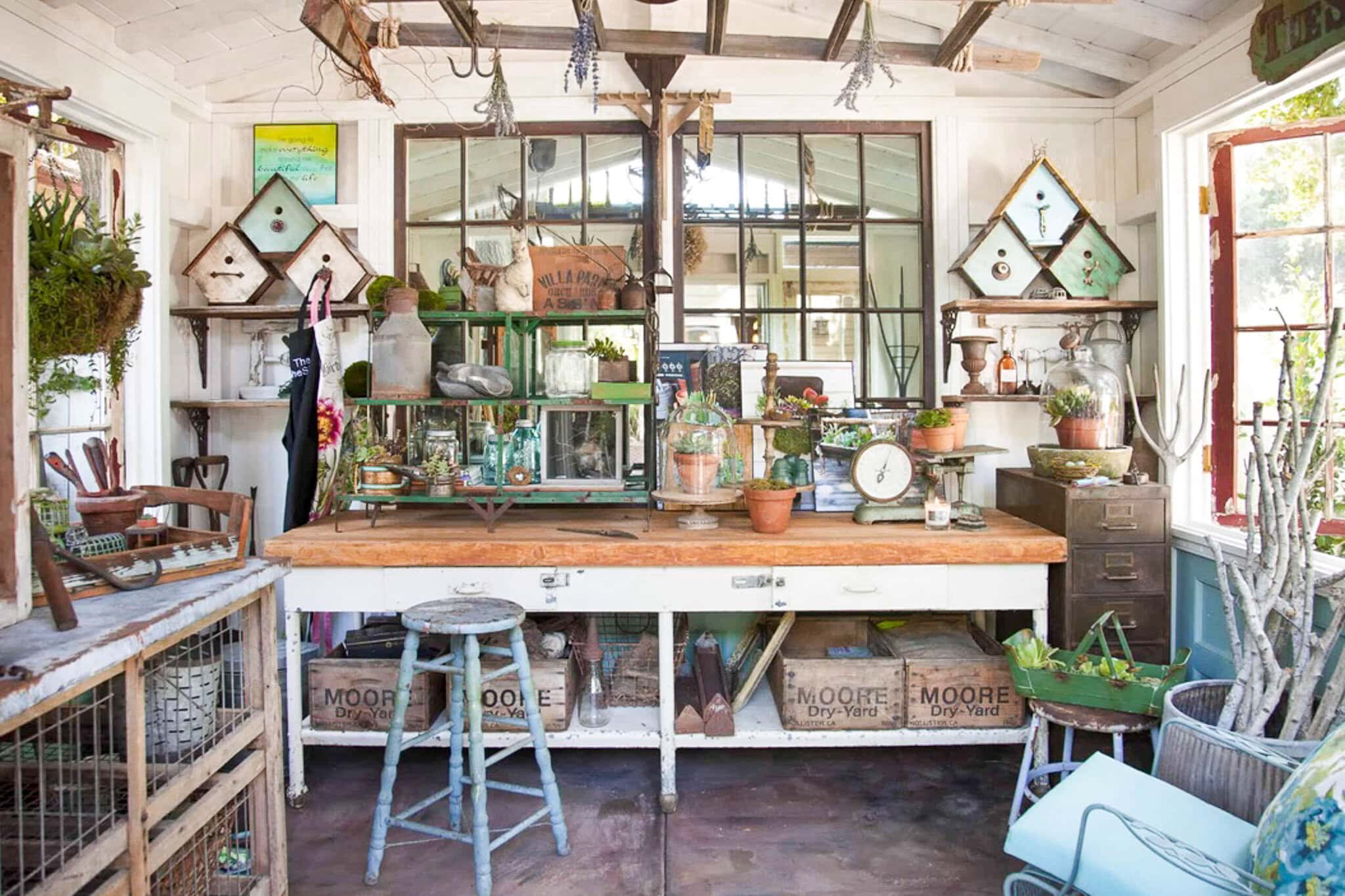 A rustic, sunlit garden shed from She Shed Interiors features a wooden workbench, potted plants, vintage scales, glass jars, and hanging tools. Large windows and mirrors reflect the bright, airy space filled with charm.