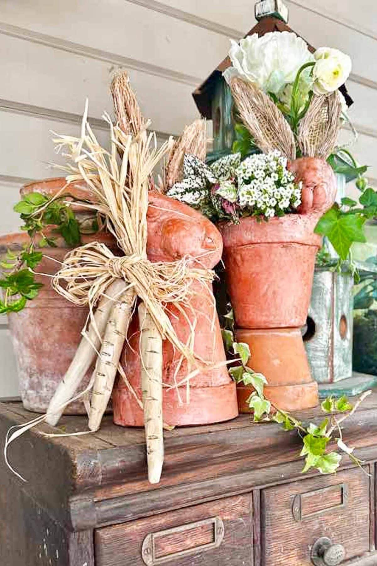 Three terracotta pots stacked to resemble bunnies with faux flowers, raffia bows, and decorative carrots—this charming Easter bunny DIY sits on a rustic wooden chest with greenery and a birdhouse in the background.