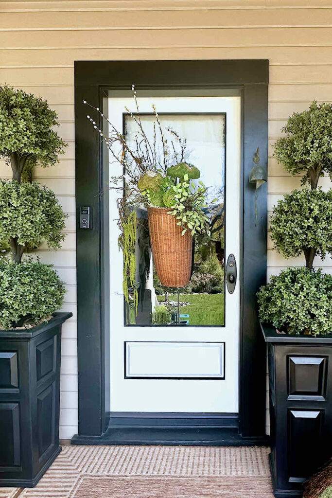 basket hanging on glass front door with two three tiered topiaries on either side of the door. Basket has faux greenery and moss mushrooms