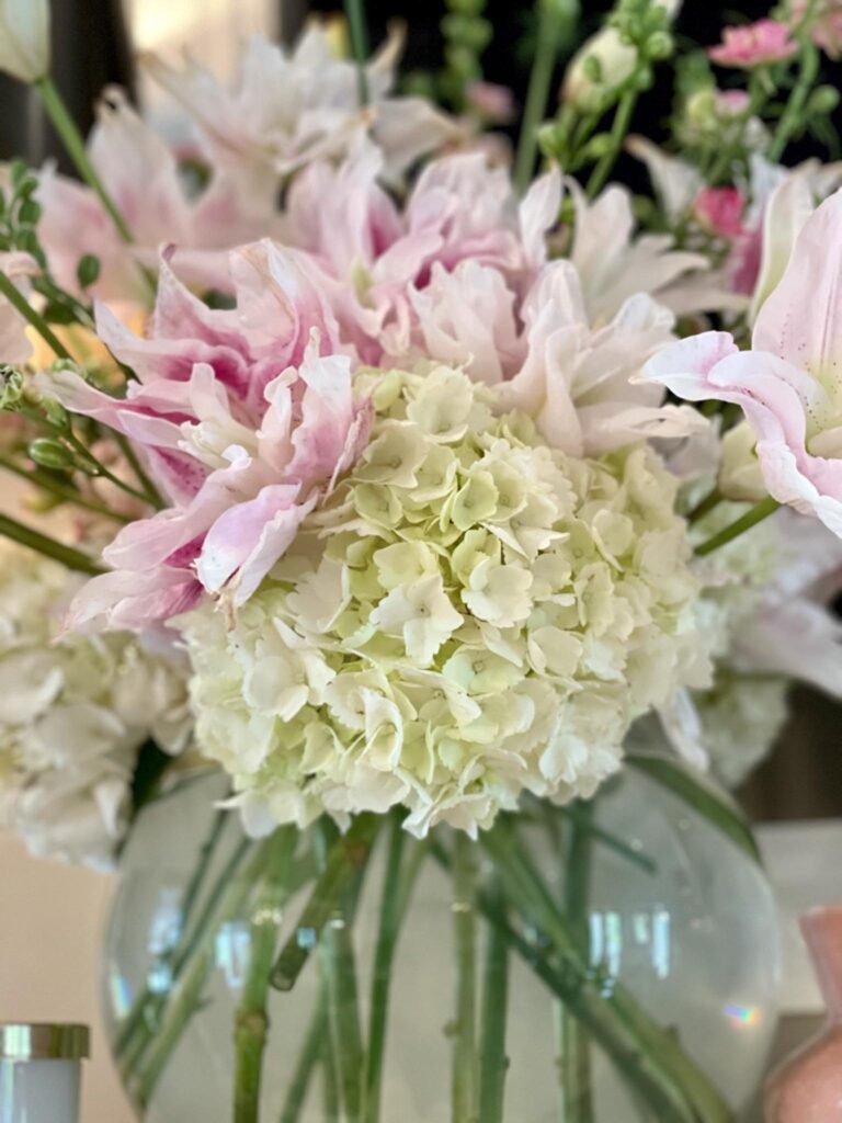 A close-up of a floral arrangement in a clear glass vase, featuring white hydrangeas and soft pink lilies. The flowers are fresh and the stems are visible through the transparent vase. The background is softly blurred, focusing attention on the beautiful bouquet.