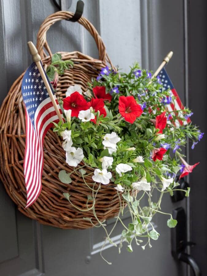 A round wicker basket hangs on a door, filled with red, white, and purple flowers. Two small American flags are placed among the flowers, creating one of the most unique wreaths for front door patriotic displays.