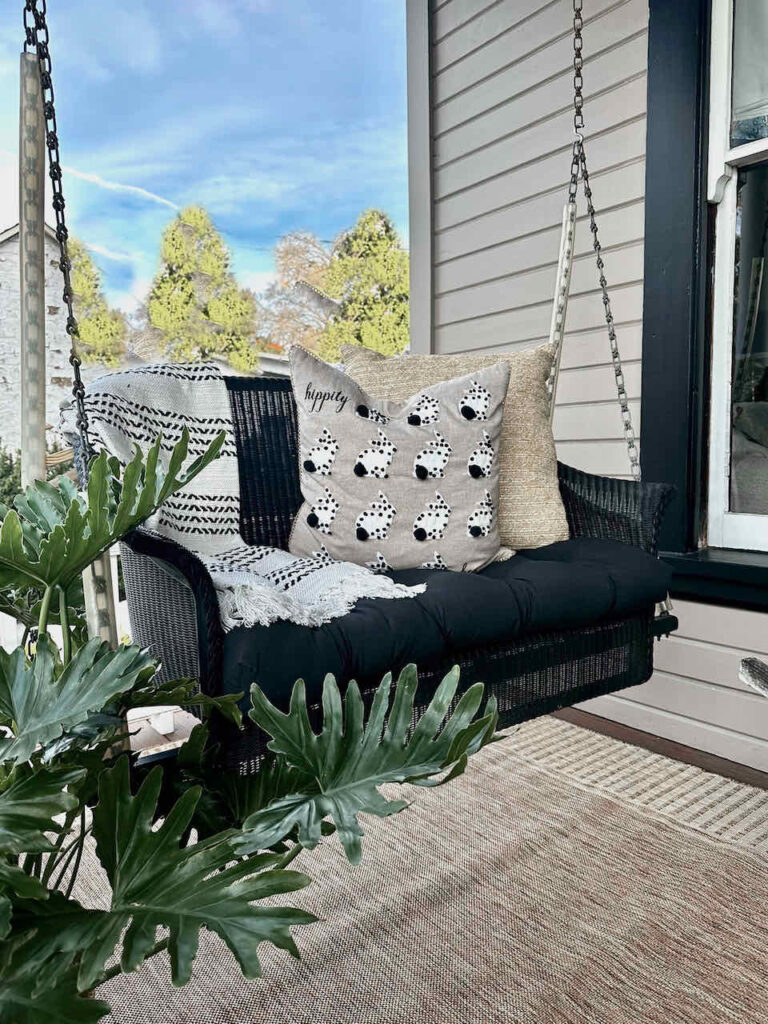 DIY spring front porch decor-Black wicker porch swing with a black and white throw blanket and two natural pillows. One pillow has tiny white bunnies with black tails decorating it—a pot of fresh greenery stands on the floor next to the swing.