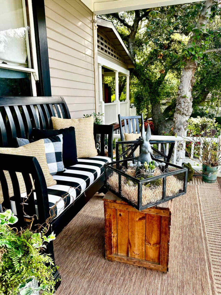 DIY spring front porch decor-A wood bench in black sits on the porch with a black and white cushion. A vintage wood trunk for a coffee table decorated with a small black cage with a cement rabbit and moss easter eggs. A black rocking chair sits in the background.