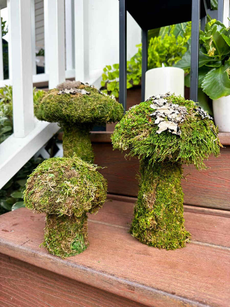 DIY spring front porch decor: A front porch with a wooden wheel barrel, birdhouse, and fresh greenery. A three-tiered topiary is next to the front door. -Three moss mushrooms sitting on the porch steps with a lantern behind them.