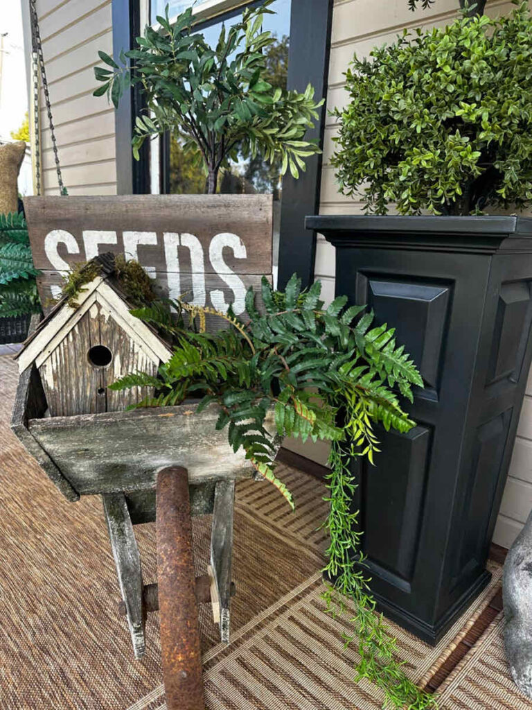 DIY spring front porch decor-Vintage wood wheel barrel filled with faux greens, a birdhouse, a small olive tree, and a seeds sign.