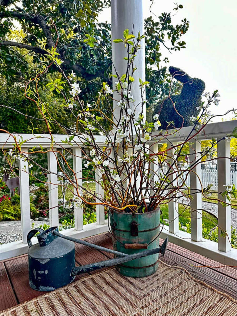 DIY spring front porch decor--A green wooden bucket filled with curly willow branches on the porch floor. A vintage green watering can sit next to the arrangement with a moss rabbit on the porch's railing.