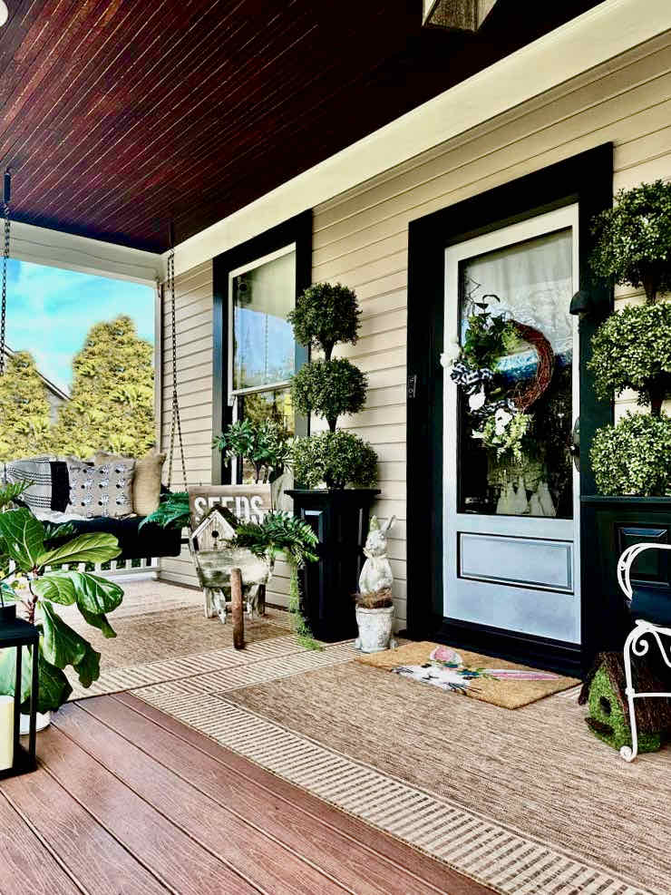 DIY spring front porch decor- Front porch of an old home with a black porch swing. A wheel barrel full of greens, a birdhouse, and a seeds sign. Two topiaries frame the glass front door with a wreath on it. It is a large carpet of natural color with a d oormat with a rabbit on it.