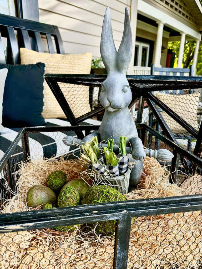DIY spring front porch decor-cement rabbit sitting in a black cage with moss Easter eggs on the porch's coffee table.