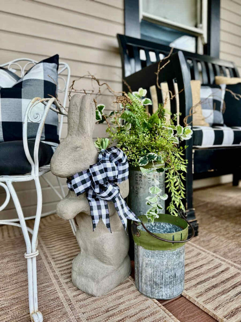 DIY spring front porch decor-Cement rabbit w ith black and white bow around his neck sitting next to a chair. two buckets of fresh greenery and twigs.