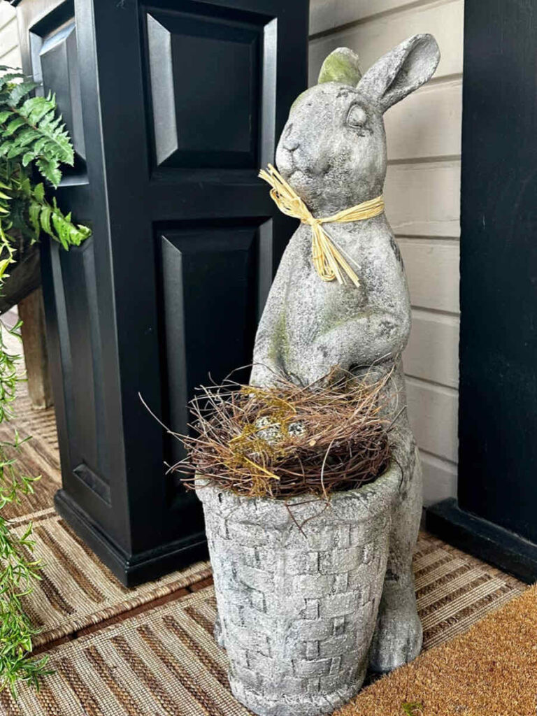A tall cement rabbit sits next to a tall black pot on the front porch, next to the front door.