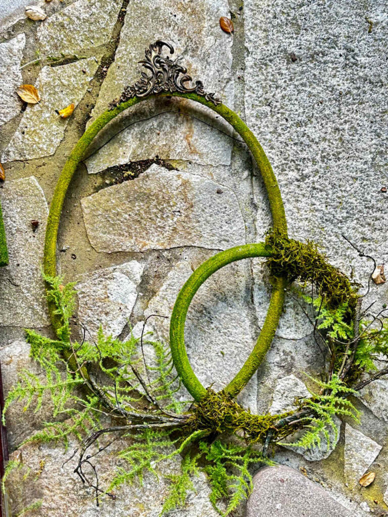 How to Make An Easy DIY Moss Frame Wall Art Gallery-the larger oval frame with the smaller round frame wired ont it. Both frames are covered in moss and decorated with faux ferns. 