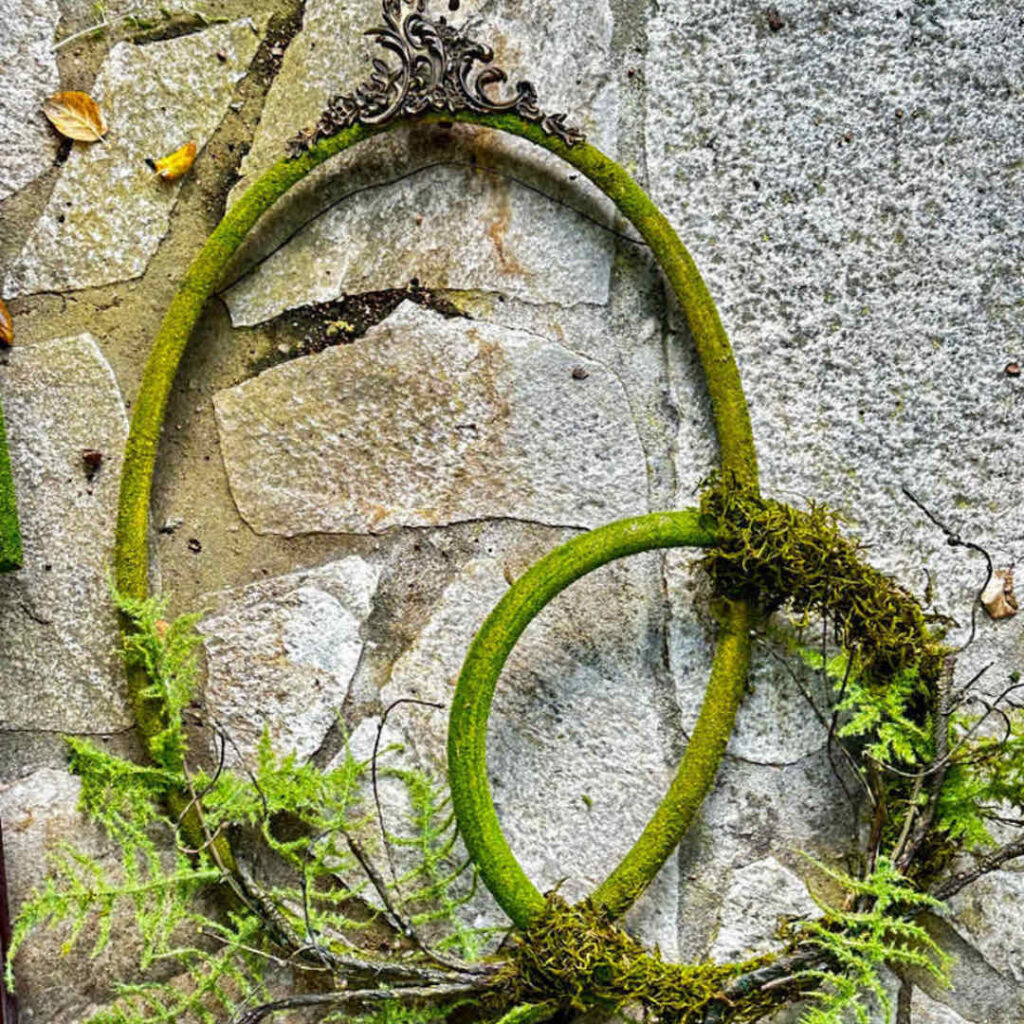 Two moss-covered vintage picture frames, one oval and one circular, are intertwined against a stone wall with ferns growing around them. The frames have ornate metallic details at the top and bottom, creating a unique summer front door display that blends nature with antique elements.