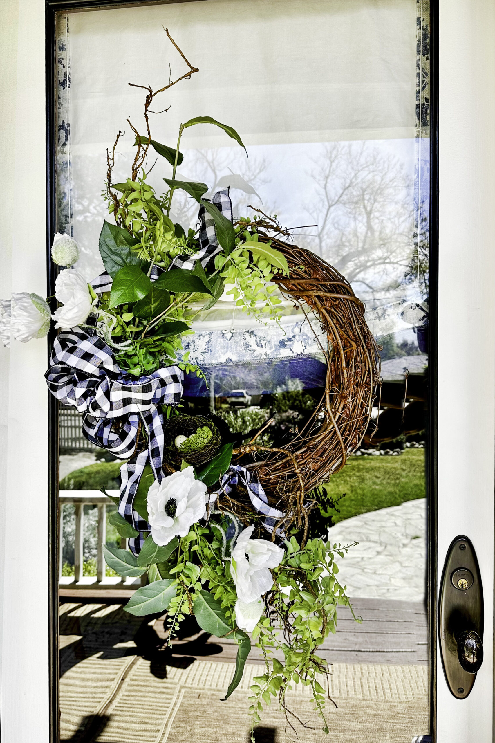 A unique summer front door showcases a grapevine wreath adorned with white flowers, green leafy accents, and a black-and-white checkered ribbon hanging on a glass door. The reflection reveals a porch and a grassy backyard with trees. A brass door handle is visible on the right side.
