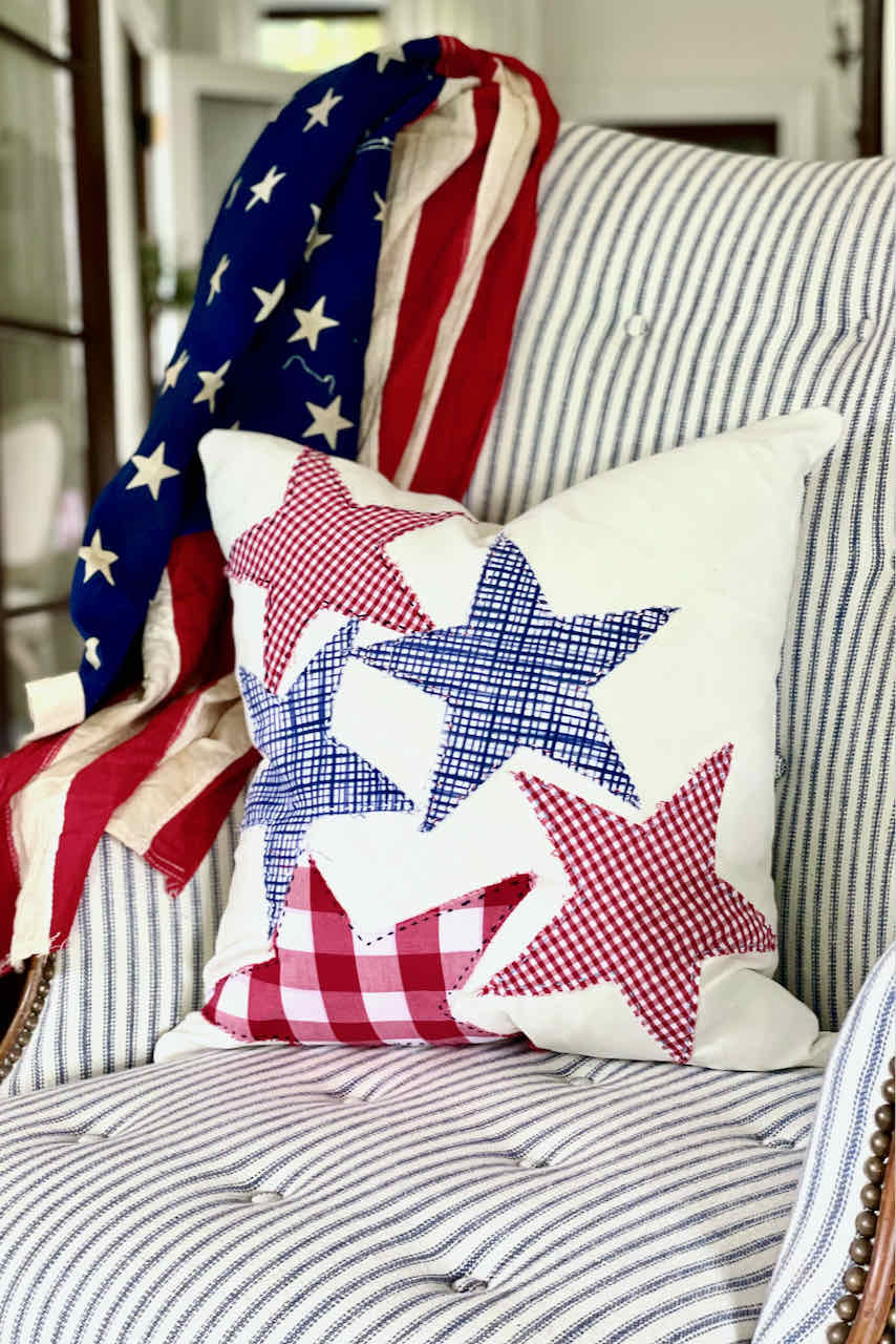 A patriotic-themed chair is draped with an American flag. A decorative pillow with red, white, and blue plaid and checkered stars is placed on the chair, which is upholstered with blue and white stripes. Perfect for Memorial Day party ideas, this setup radiates a festive and cozy ambiance.