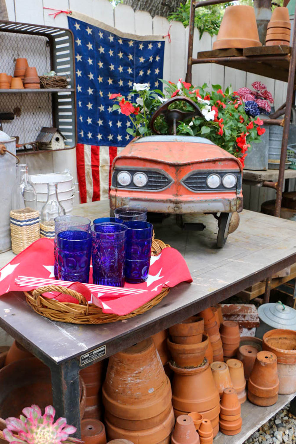 Garden shed table with old vintage car decorated with red and white impatients. Shelf lined with terra cotta pots. A tray of bright blue glasses is sitting on top with a flag hanging on the fence.