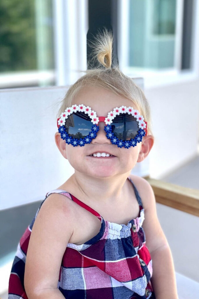 Little girl in patriotic sunglasses that are red, white and blue and the lenses are surrounded with small flowers