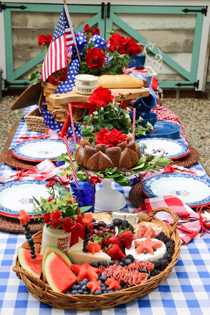 The table was decorated for the party with red, white, and blue. Checkered tablecloth with a picnic basket filled with red geraniums. Charcuterie board, cake, and a loaf of bread on the table for dinner.. a pile of enamel dishes sitting on the table.