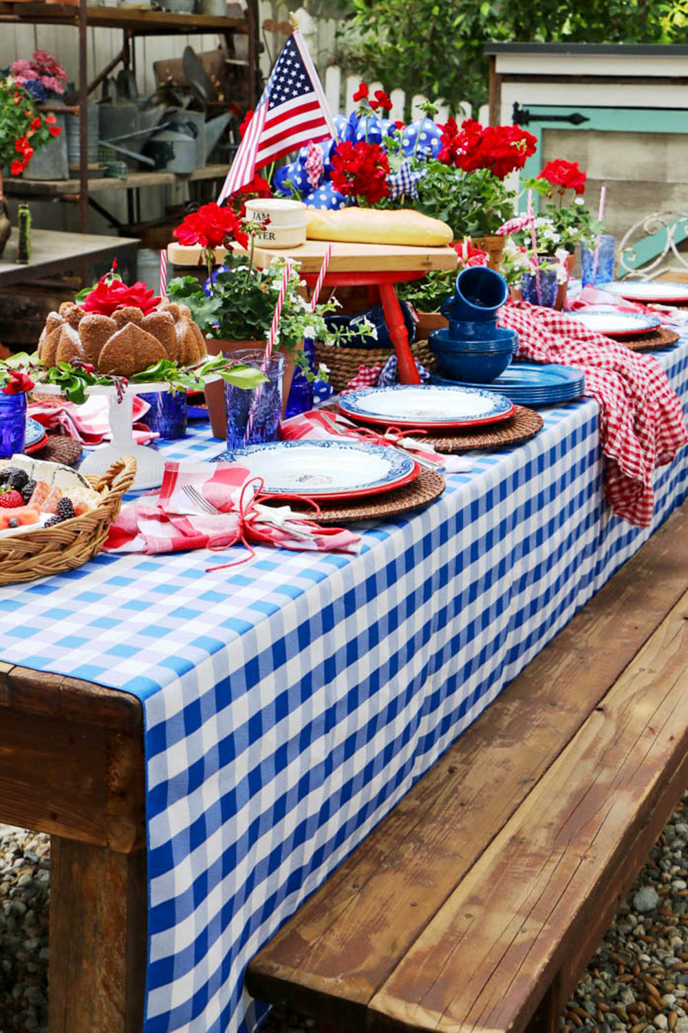 The table was decorated for the party with red, white, and blue, and a checkered tablecloth with a picnic basket filled with red geraniums. Flags are hanging on the fence for decoration. A pile of enamel dishes is sitting on the table.