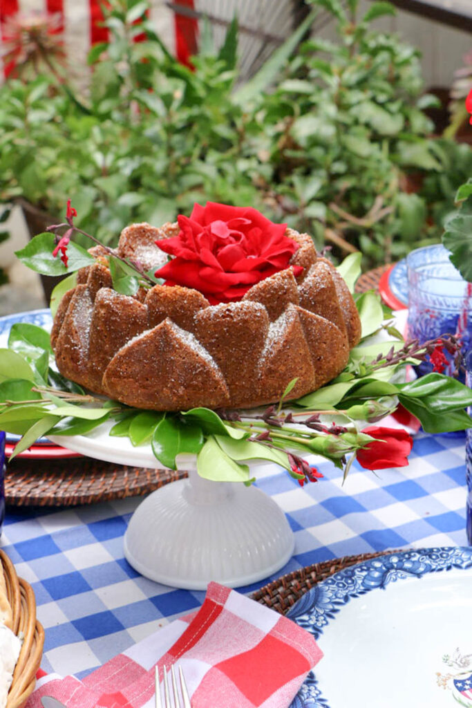 Memorial Day Party Ideas- Sherry Bundt cake displayed on a white glass cake plate. The cake has fresh greenery around the edge and a new red rose.