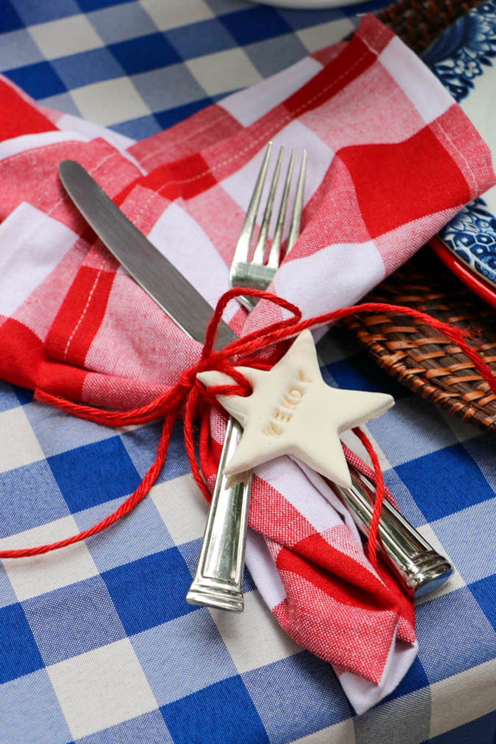 Silverware lying on a blue and white check tablecloth with red and white check napkins. Silverware is wrapped up with a cut-out white star that has a name imprinted in the star.