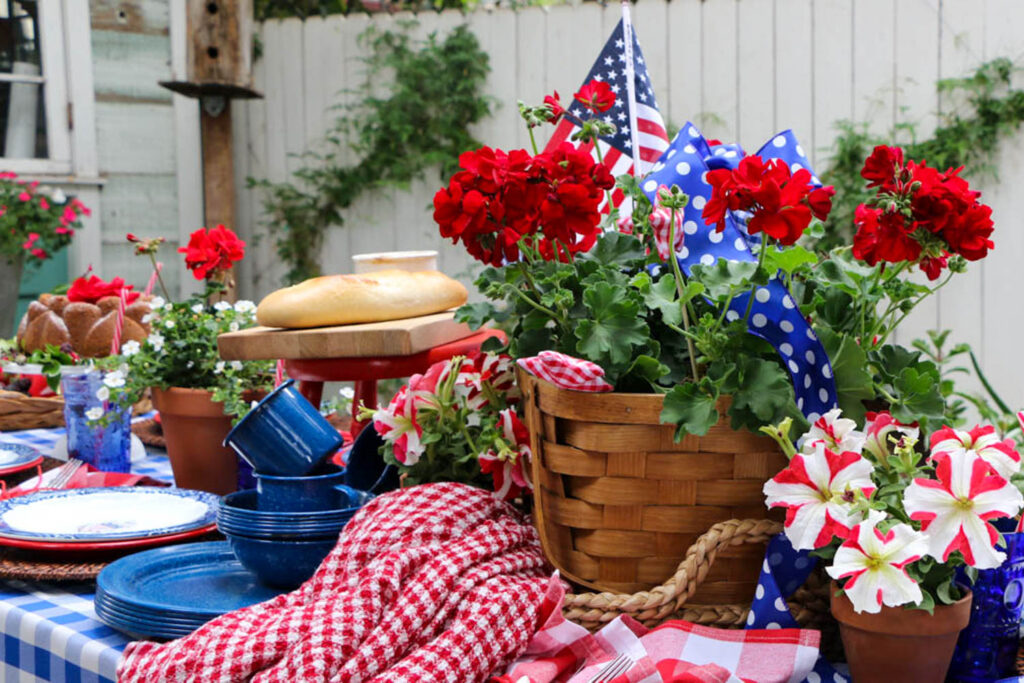 Memorial Day Party Ideas- Table decorated for party with red white and blue. Checkered table cloth with a picnic basket filled with red geraniums.
