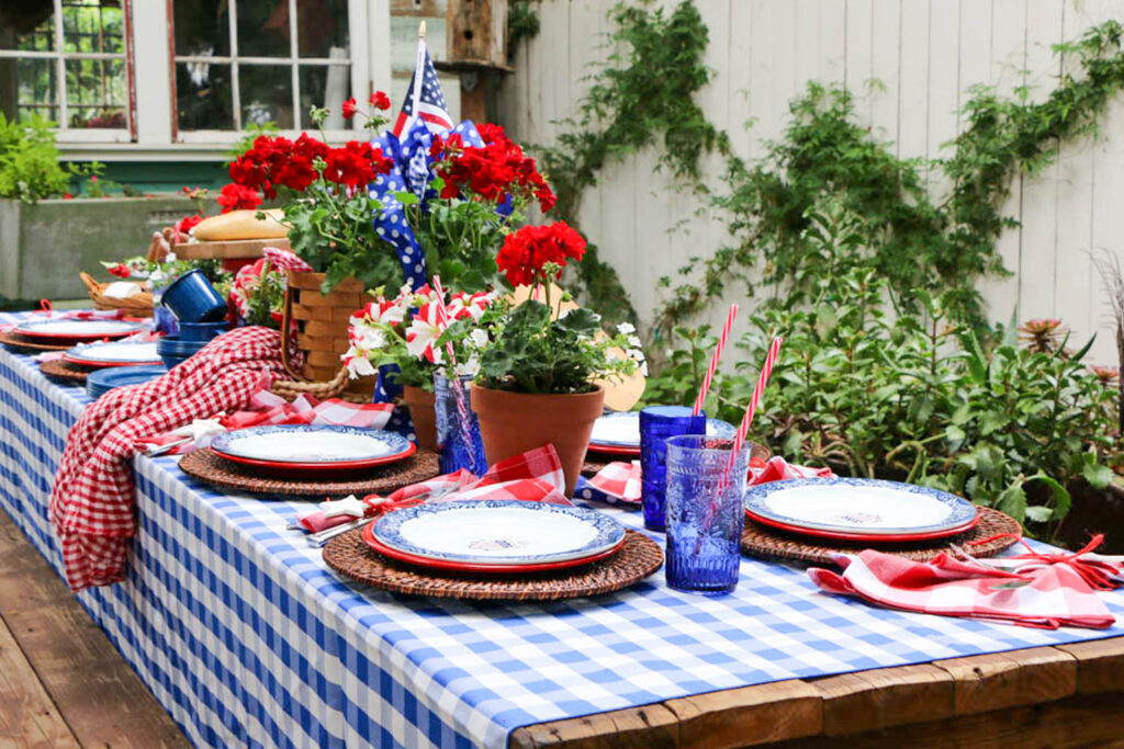 Memorial Day Party Ideas-Table decorated for a red, white, and blue party. Checkered tablecloth with a picnic basket filled with red geraniums. Pots of red and white striped petunias, Bright blue glasses, and patriotic plates.