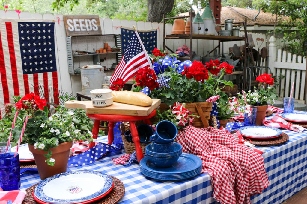 Memorial Day Party Ideas-Table decorated for a party with red white and blue. Checkered table cloth with a picnic basket filled with red geraniums. Flags hanging on the fence for decoration. pile of enamel dishes sitting on the table.