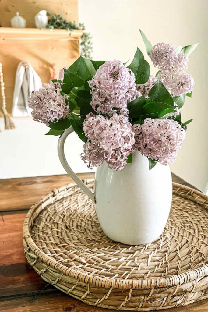 A WHITE PITCHER FILLED WITH PURPLE LILACS SITTING IN A WICKER TRAY