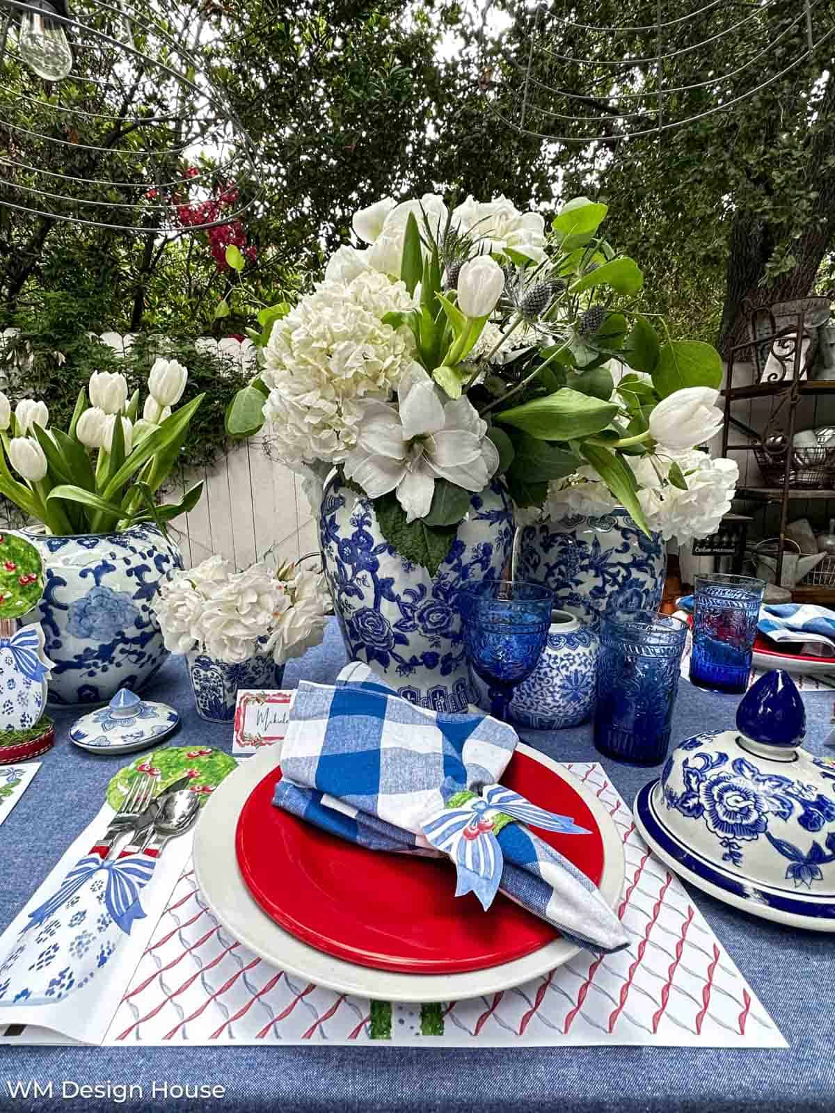 4th of July table setting with red and white plates, blue plaid napkins, and a white floral centerpiece in a blue and white vase