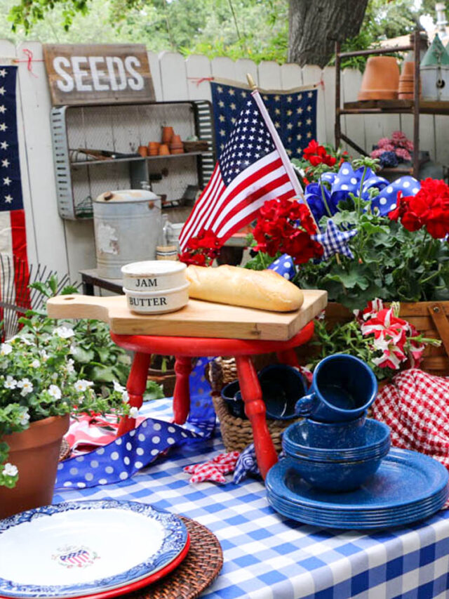Memorial Day Party Ideas-Table decorated for a party with red white and blue. Checkered table cloth with a picnic basket filled with red geraniums. Flags hanging on the fence for decoration. pile of enamel dishes sitting on the table.