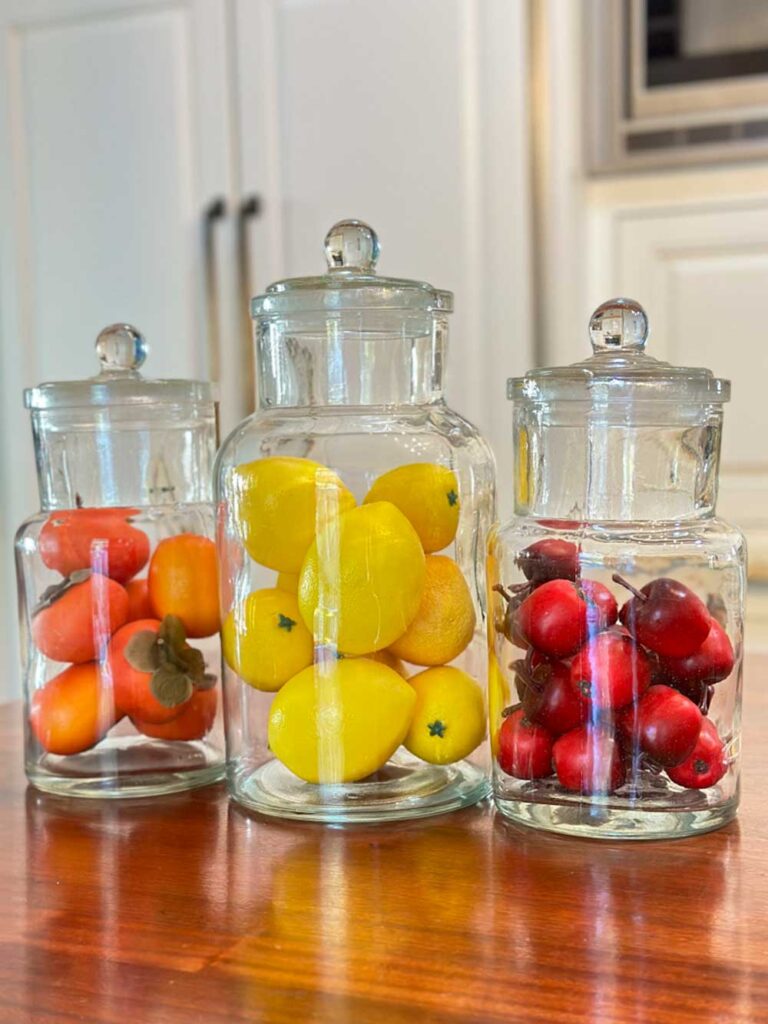 three apothecary jars on the kitchen counter filled iwth fruit. One has persimmons, one has lemons and the third jar has crab apples. 