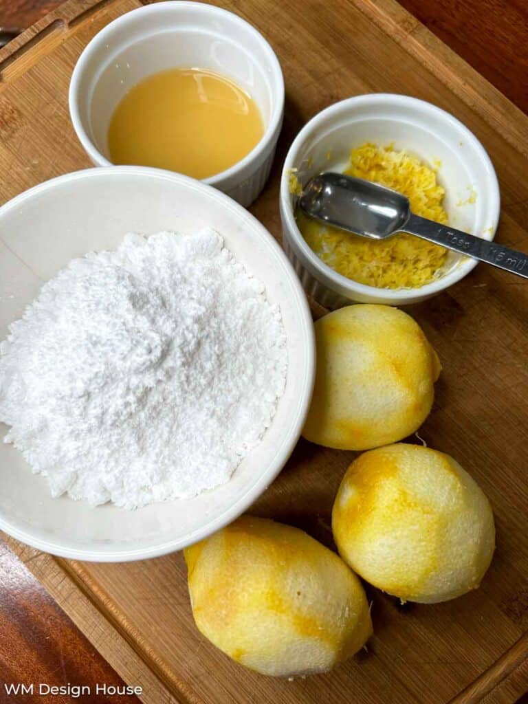 A wooden cutting board with ingredients for a recipe. Visible items include three whole lemons, a bowl of powdered sugar, a bowl of lemon juice, a bowl containing lemon zest, and a tablespoon measure.