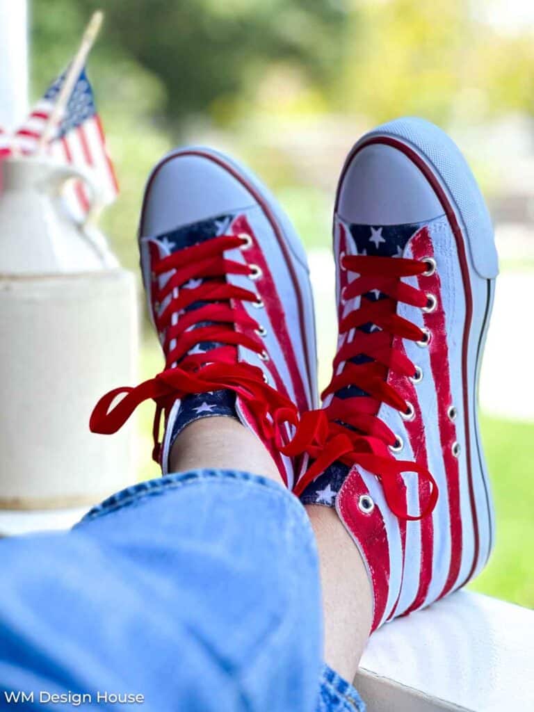 feet up on a railing with patriotic tennis shoes resitng on the rialing. The shoes have red and white stripes with a blue tongue that has white stars. Laces are red velvet