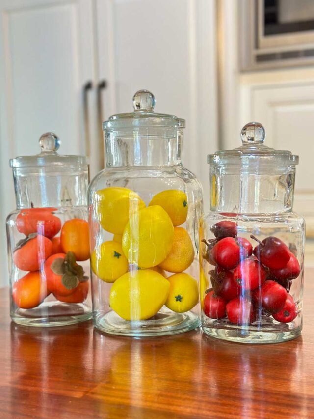 three apothecary jars on the kitchen counter filled iwth fruit. One has persimmons, one has lemons and the third jar has crab apples.