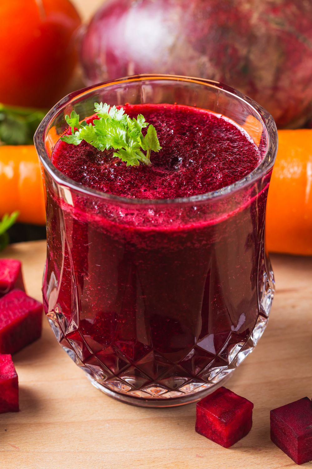A glass of dark red beetroot juice garnished with fresh parsley, surrounded by raw beets, a carrot, a tomato, and diced beet cubes on a wooden surface—perfect for those curious about how long do cooked beets last.
