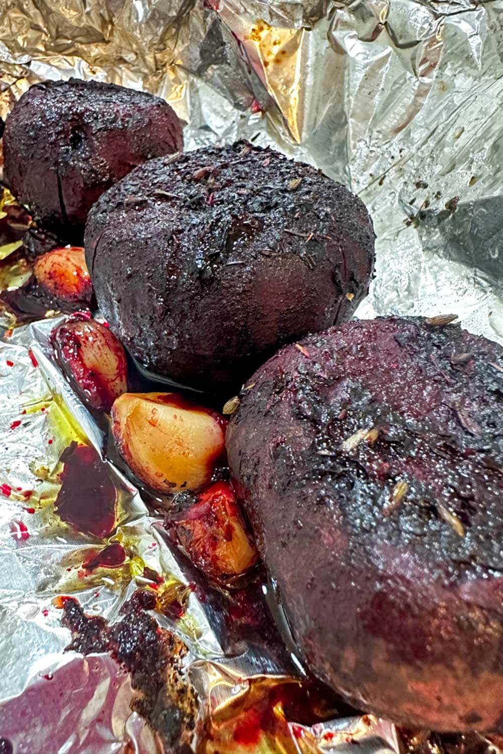 Three whole roasted beets with garlic cloves rest on crinkled aluminum foil, lightly charred and glistening with juices—perfect for enjoying now or storing in the fridge if you’re wondering how long do cooked beets last.