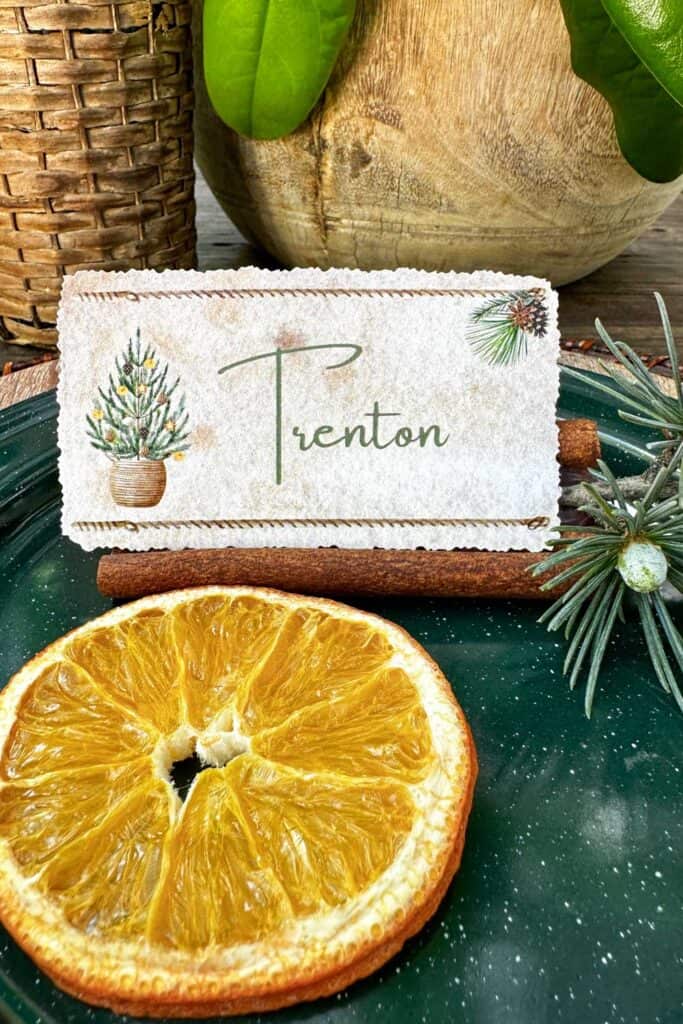 A name place card holder made of sweet smelling cinnamon sticks, fresh greens and a slice of dried orange.