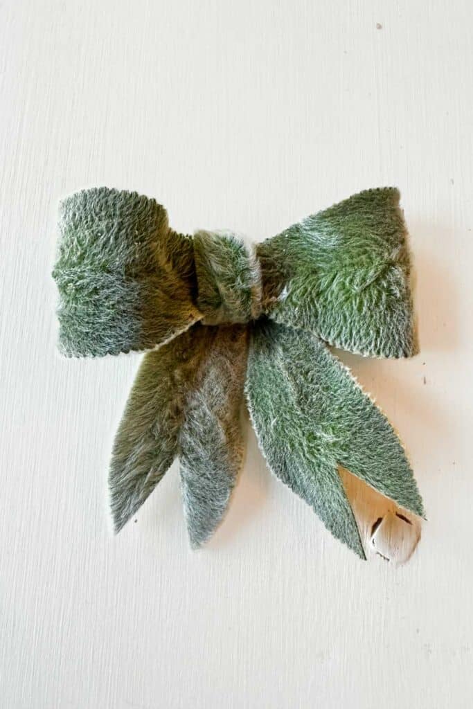 A bow made of soft, fuzzy green material resembling lamb's ear plant leaves, tied in the center with additional leaf-like extensions hanging down. The background is a smooth, off-white surface.