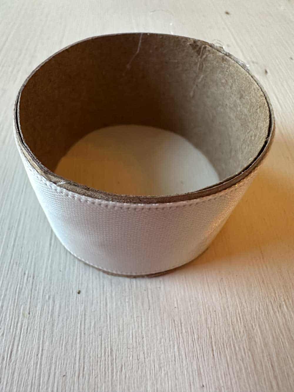 A plain, empty cardboard ring with a white exterior surface is standing on a light-colored wooden table. The ring appears to be part of a roll, possibly for ribbon or tape, with the inner part hollow.