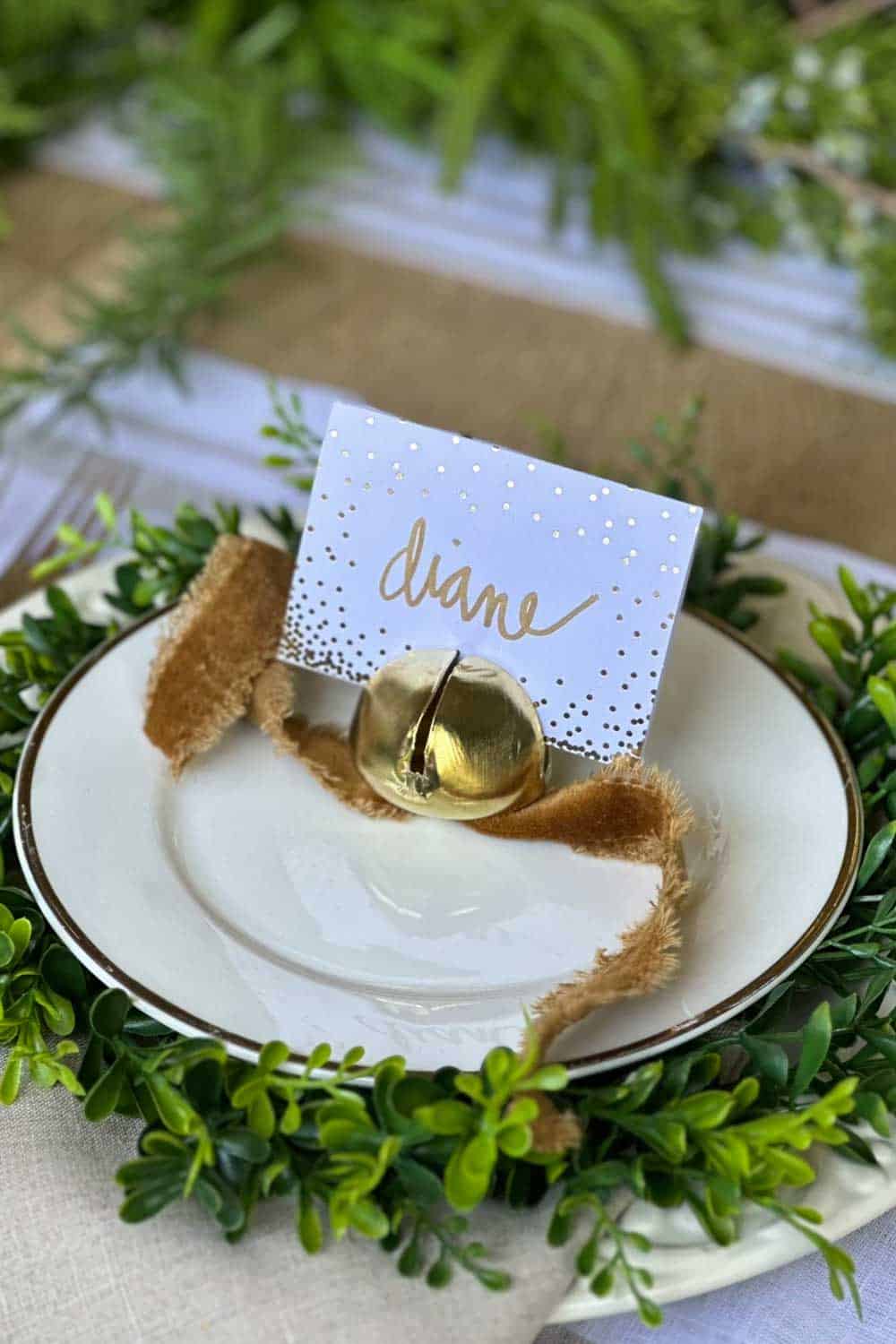 A whte plate with a gold bell and goldd velvet ribbon holding a place card for Christmas dinner. 