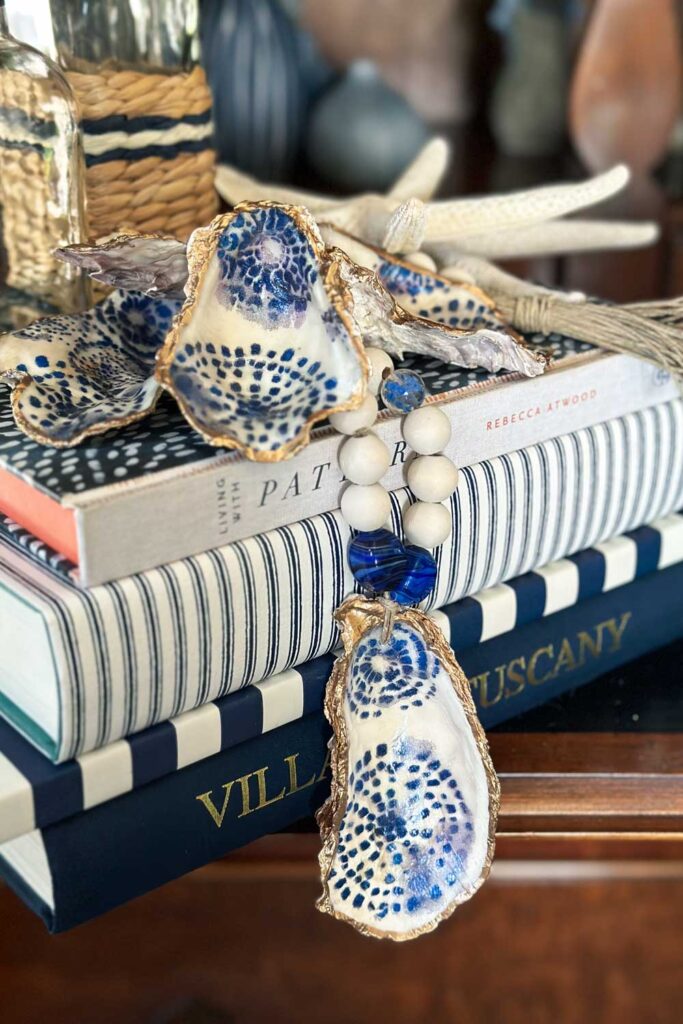 wood beaded garland with oyster shell. A wood garland with a decoupaged oyster shell hanging over a few books for decoration in a living room that is all blue and white.