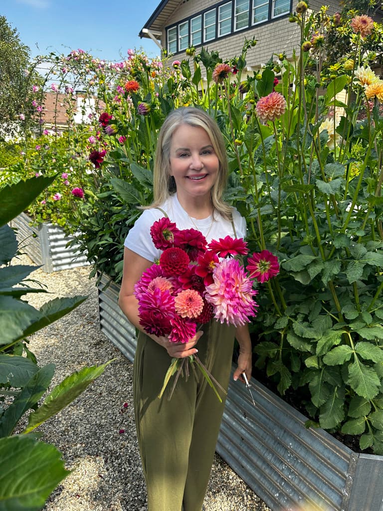 A woman stands in a garden holding vibrant pink and red flowers. She is dressed in a white t-shirt and green pants. Bright green plants and colorful flowers surround her, with a building visible in the background. The sky above is clear and blue.