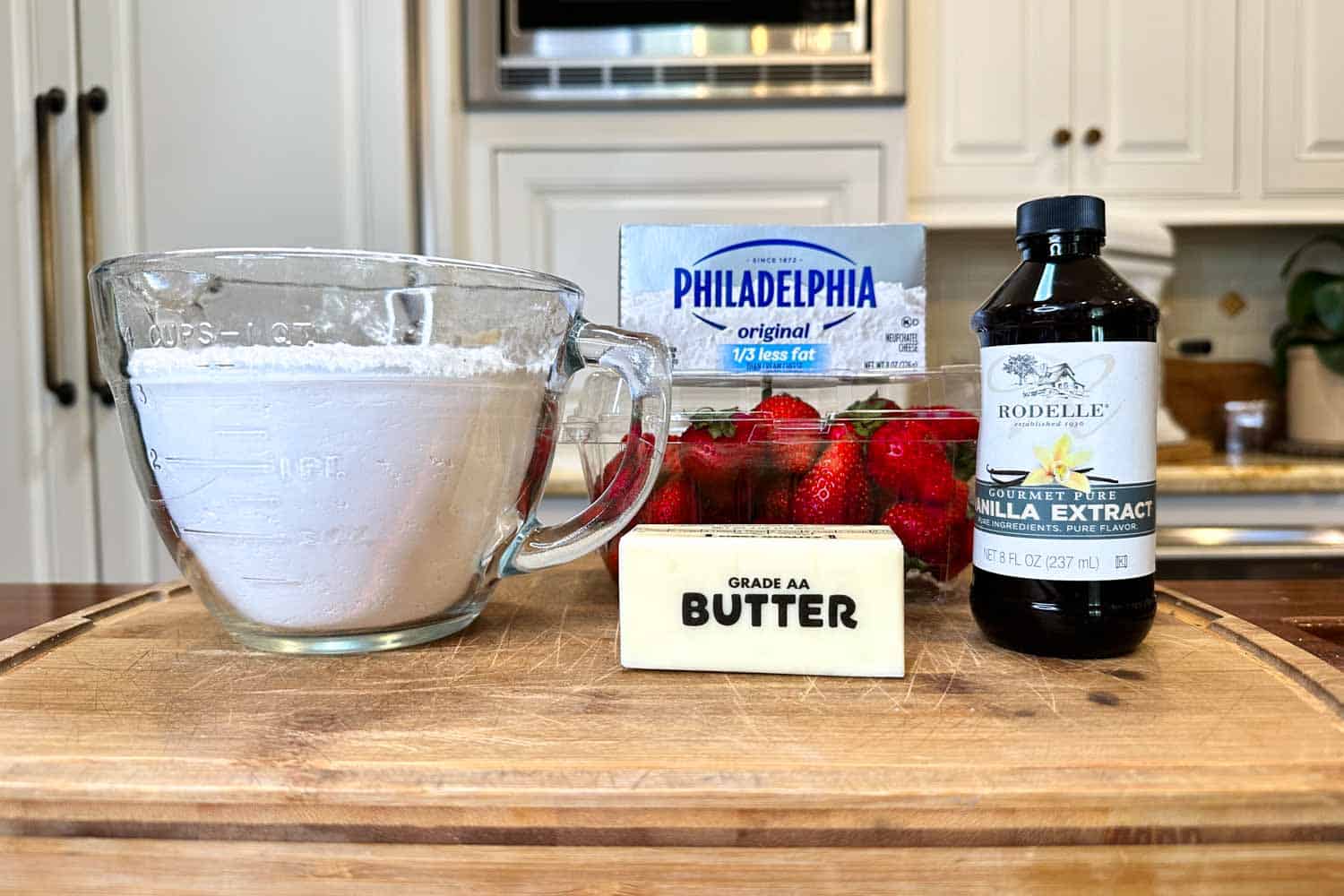 A wooden kitchen countertop with various ingredients: a glass measuring cup filled with powdered sugar, a package of Philadelphia cream cheese, a bottle of Rodelle vanilla extract, a stick of Grade AA butter, and a carton of fresh strawberries in the background.