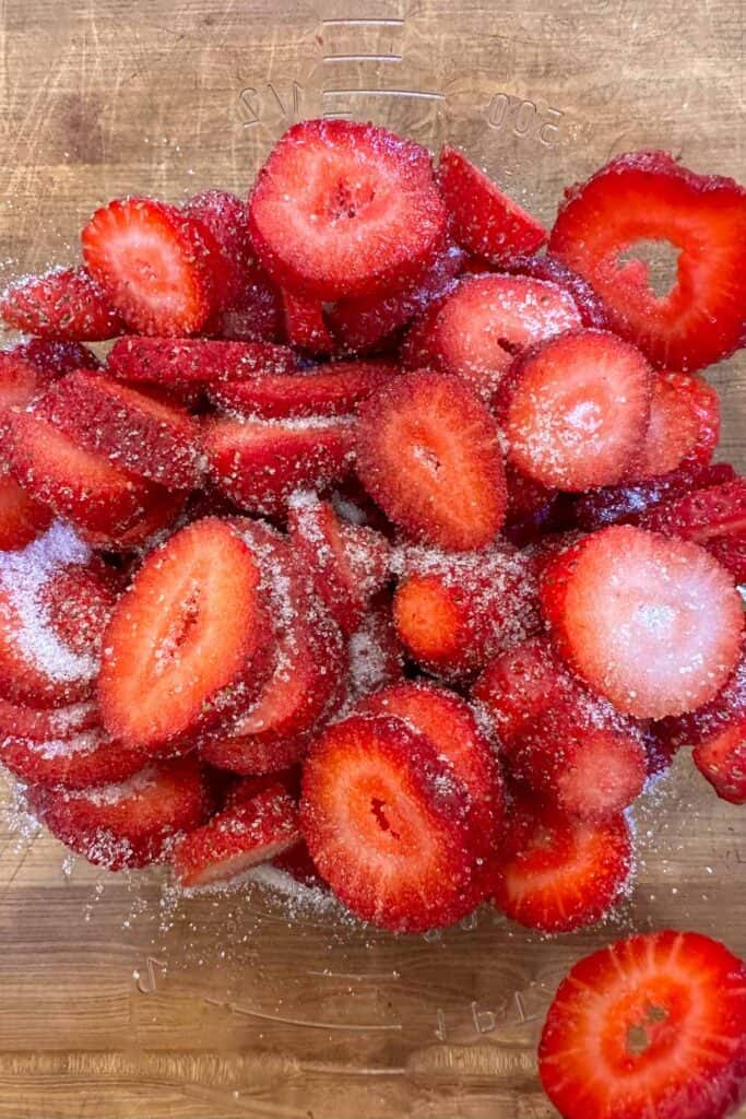 A pile of freshly sliced strawberries sprinkled with sugar is placed on a wooden surface. The strawberries are vibrant red, with the sugar glistening on top, creating a contrast between the textures.