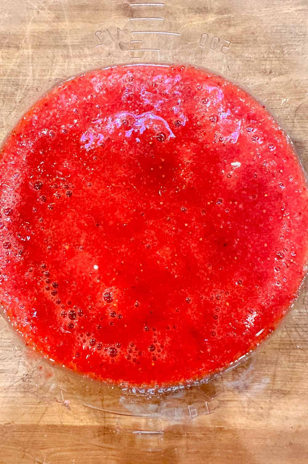 A clear measuring jug filled with vibrant red strawberry puree, placed on a wooden surface. The puree is smooth with some bubbles on the surface, indicating it has been freshly blended or mixed.
