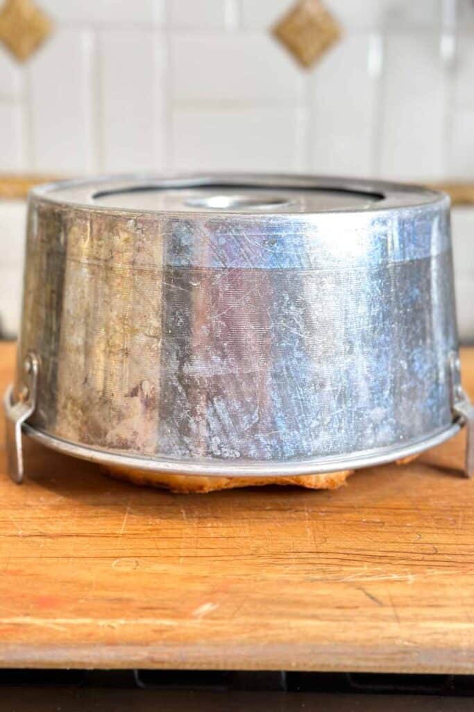 A metal cake pan is turned upside down on a wooden surface. The baked cake inside is visible through the bottom of the pan. The background features a tiled wall with some diamond-shaped accent tiles.